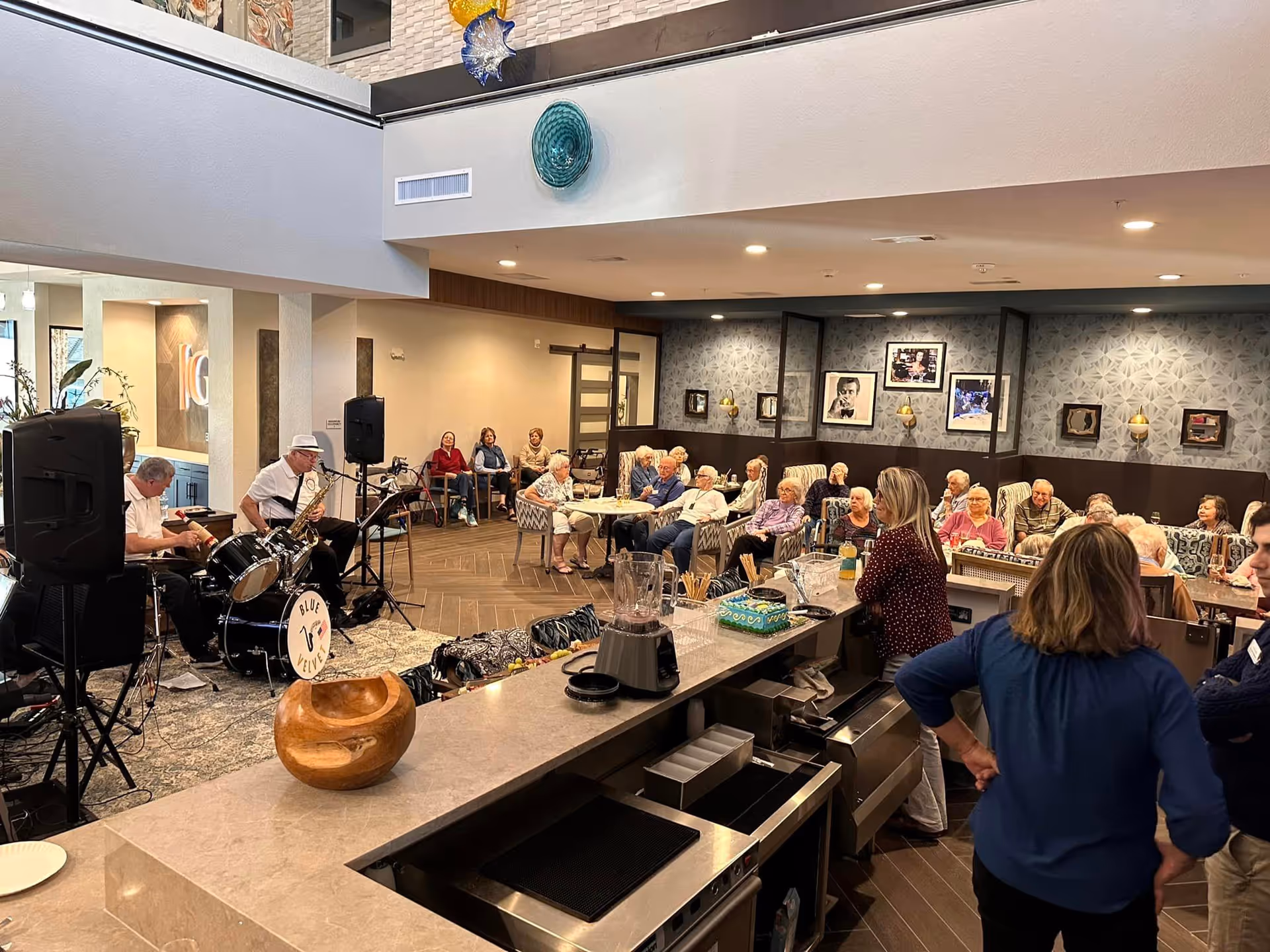 A lively indoor gathering at The Gallery at Port Orange featuring elderly residents seated and enjoying live music performed by two musicians, one playing drums and the other a saxophone. The room has modern decor with patterned wallpaper, framed pictures on the wall, and a bar counter in the foreground with a blender and other items. Several staff members are also present, engaging with the residents.