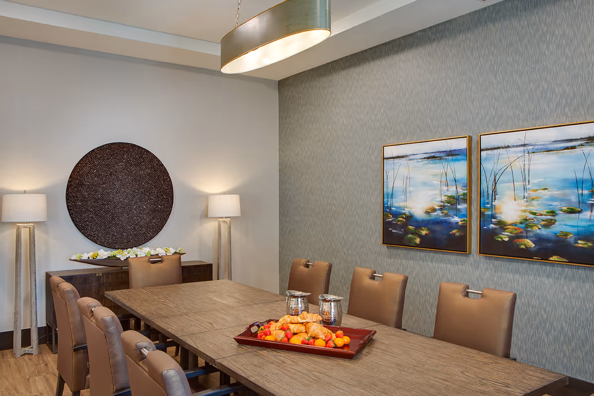 A modern dining room with a long wooden table surrounded by brown leather chairs. On the table is a red tray with croissants and assorted fruits, along with two silver pitchers. The room features two floor lamps with white shades, a large round decorative wall piece, and two framed paintings of a water scene with lily pads on a textured blue-gray wall.
