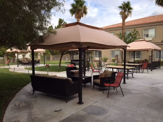Outdoor seating area with multiple canopies providing shade over chairs and tables on a concrete patio. Palm trees and a two-story building with windows are visible in the background.