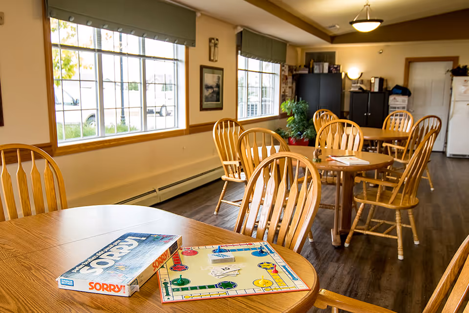 A bright room with several wooden tables and chairs arranged for group activities or dining. On the nearest table, there is a Sorry! board game set up with game pieces and cards. Large windows let in natural light, and there are plants and cabinets in the background.