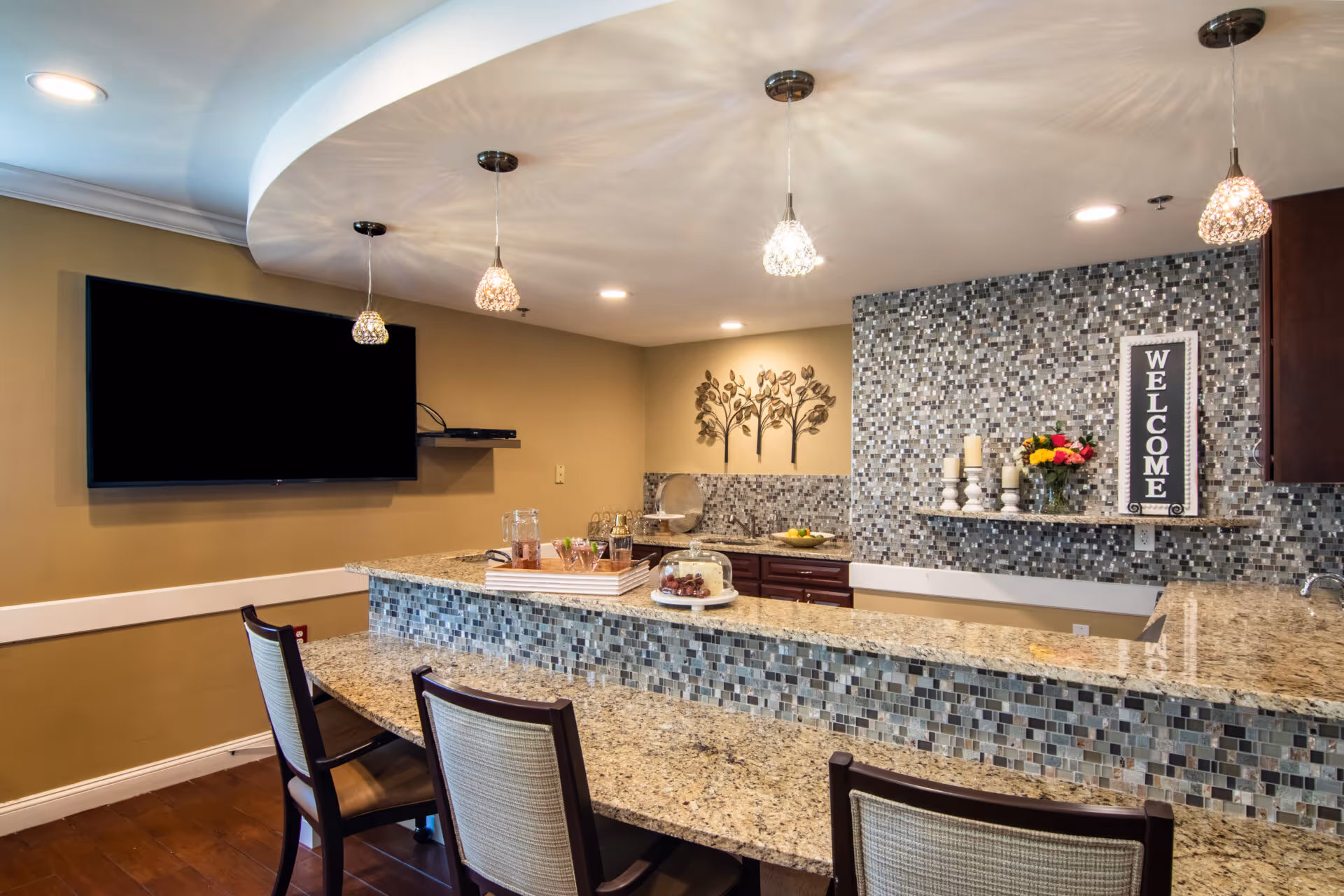 Modern communal kitchen and bar area with granite countertops, mosaic tile backsplash, pendant lights, seating, and a wall-mounted TV.