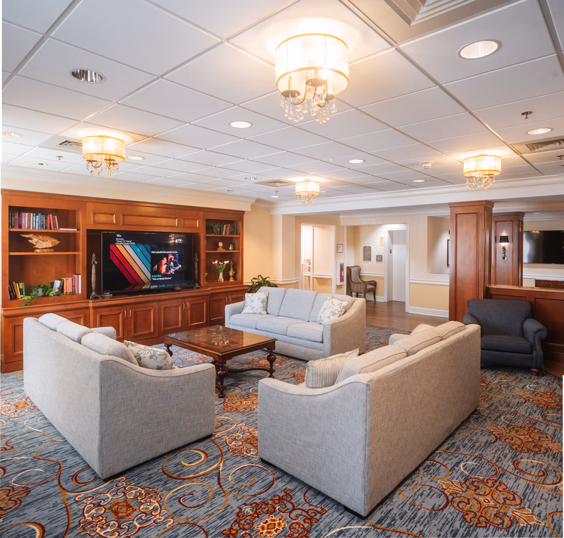 A spacious and well-lit living room area with three light gray sofas arranged around a wooden coffee table. The room features a large built-in wooden entertainment center with a flat-screen TV and shelves holding books and decorative items. The ceiling has recessed lighting and elegant hanging light fixtures. The floor is covered with a patterned carpet, and there is an armchair in the corner near a hallway.