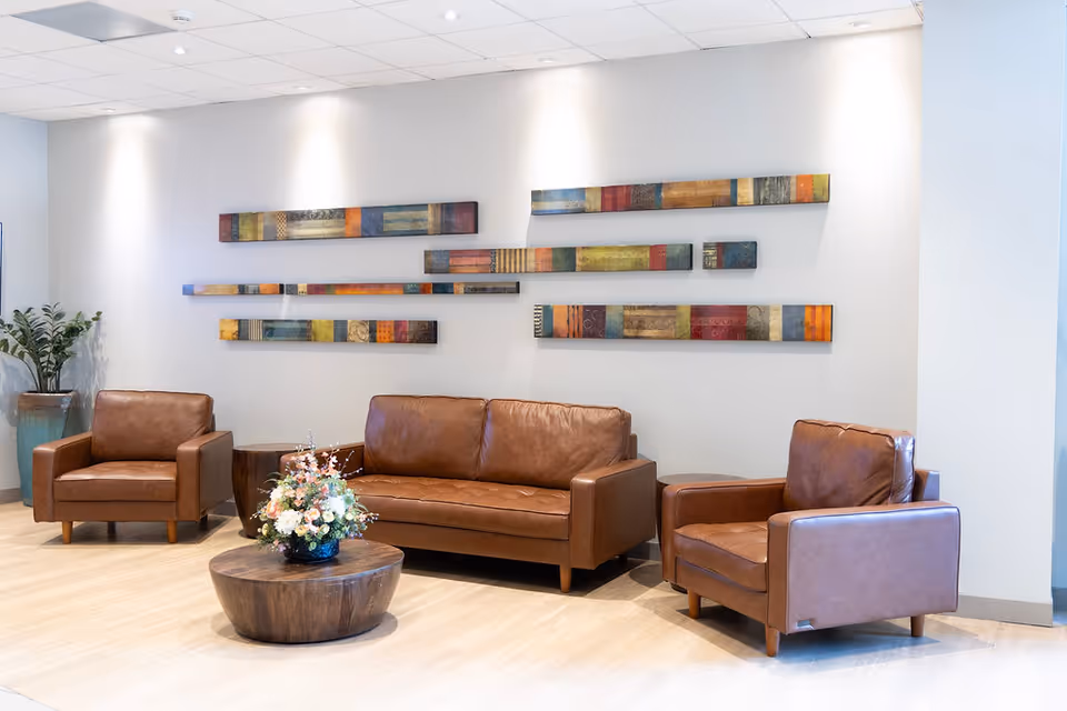 A modern lounge area with a brown leather sofa and two matching armchairs arranged around a round wooden coffee table with a floral arrangement. The wall behind features colorful rectangular abstract art pieces, and there is a large potted plant in the corner.