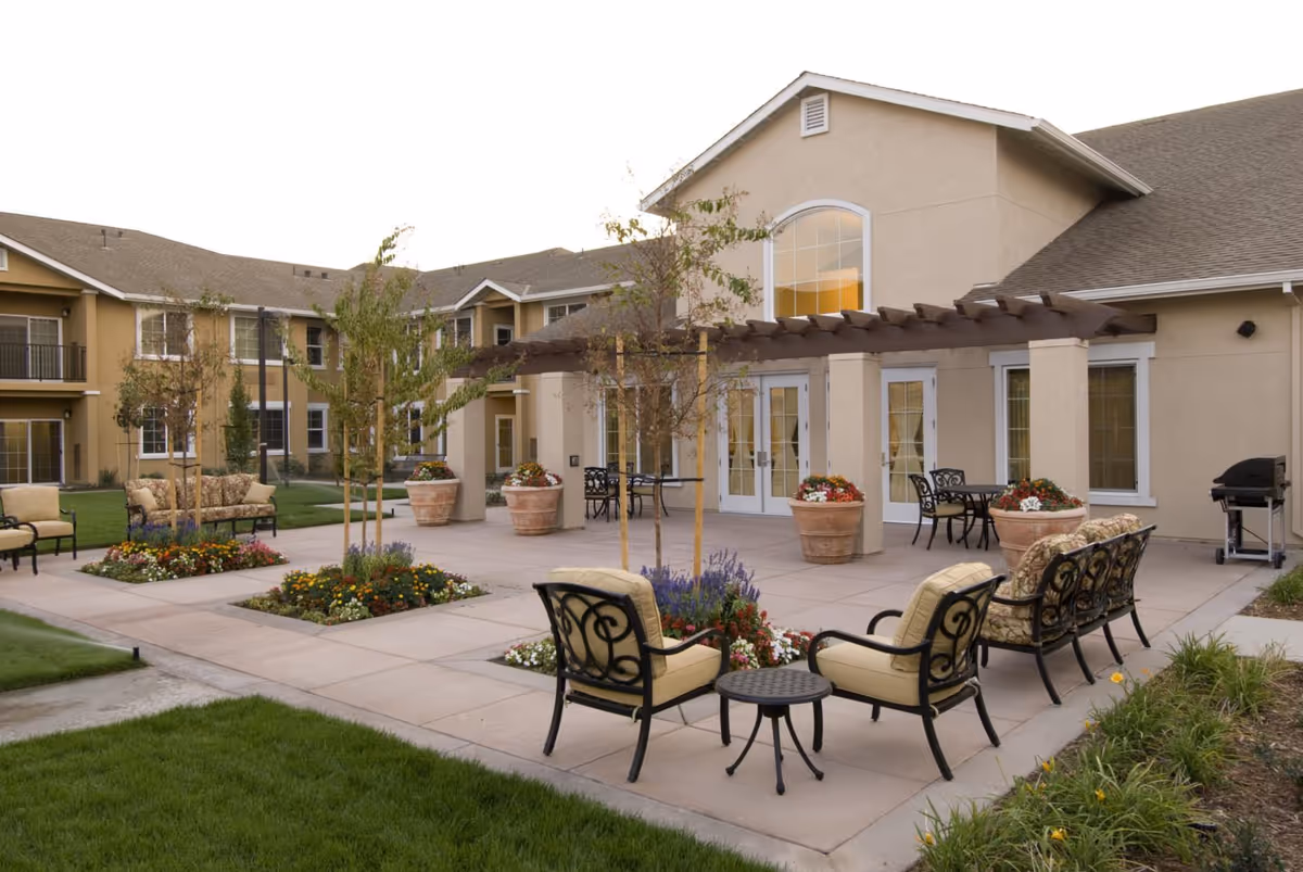 Outdoor patio area at Prestige Assisted Living at Manteca featuring cushioned chairs and sofas arranged around small tables, potted plants with flowers, young trees, and a pergola attached to the building. The building has beige walls and large windows, with a barbecue grill visible on the right side.