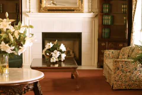 A cozy living room with a floral patterned armchair, a wooden coffee table with white flowers in a vase, a fireplace with a white mantel, a large mirror above the fireplace, a wooden bookshelf with green books, and a round table with a vase of white flowers.
