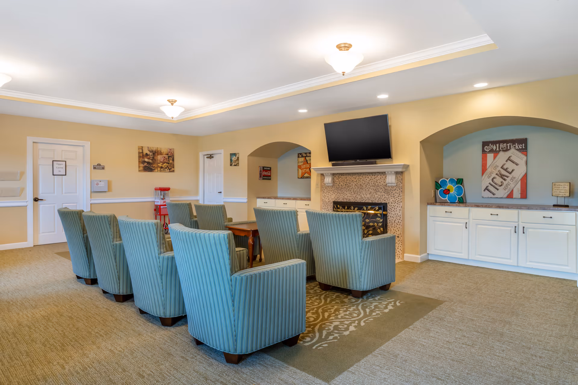 A communal lounge with upholstered armchairs arranged facing a wall-mounted TV above a fireplace.
