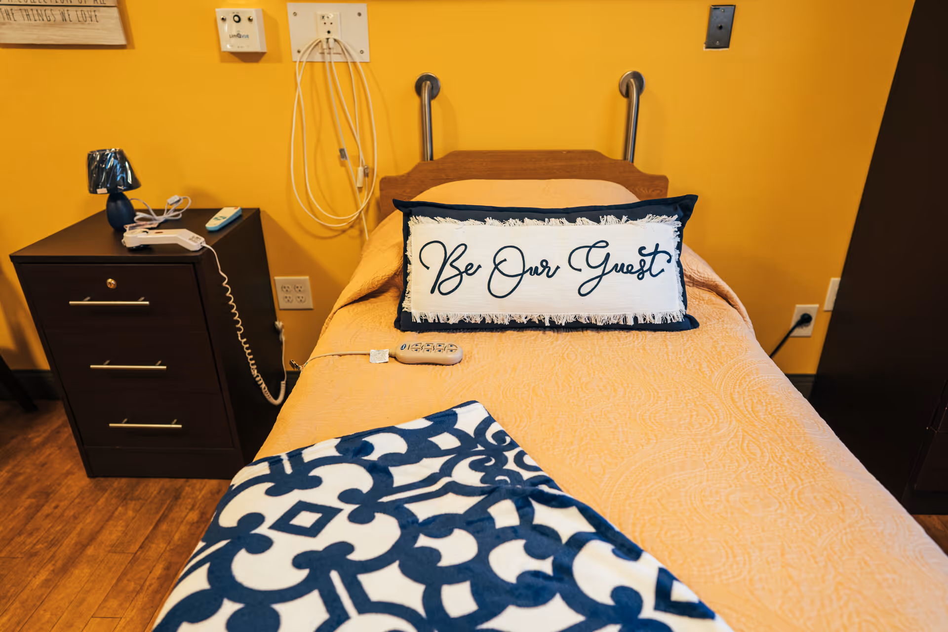 A single bed with a yellow bedspread and a decorative pillow that says 'Be Our Guest' in a room with yellow walls. Next to the bed is a dark wooden nightstand with a small lamp, a telephone, and a remote control. The floor is wooden, and there are electrical outlets and medical equipment on the wall behind the bed.