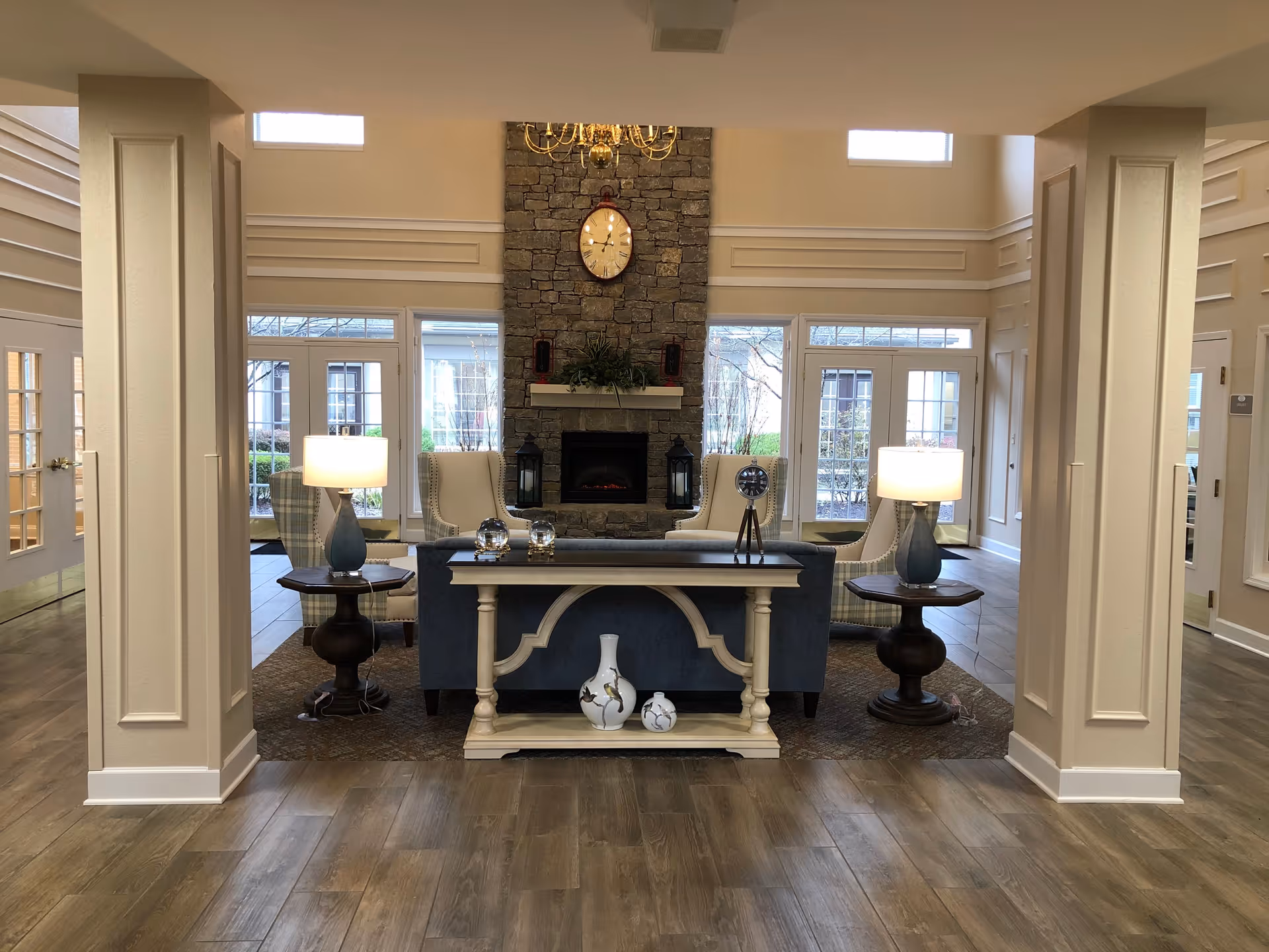 A cozy living room area with a stone fireplace centered on the back wall, flanked by two large windows. In front of the fireplace are two beige armchairs and a blue sofa with a decorative console table behind it. The console table holds two vases and two glass ornaments. There are two side tables with lamps on either side of the sofa. The room has wooden flooring and cream-colored walls with decorative molding.