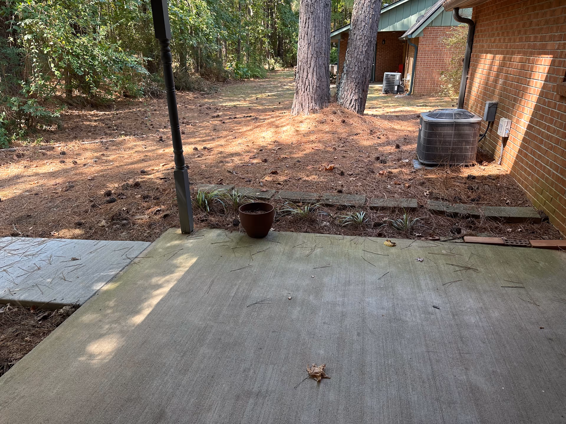 A concrete patio area with a small potted plant on the edge, adjacent to a brick building with two air conditioning units. The patio overlooks a wooded area with pine trees and a ground covered in pine needles and pine cones. A narrow concrete walkway extends from the patio into the wooded area.