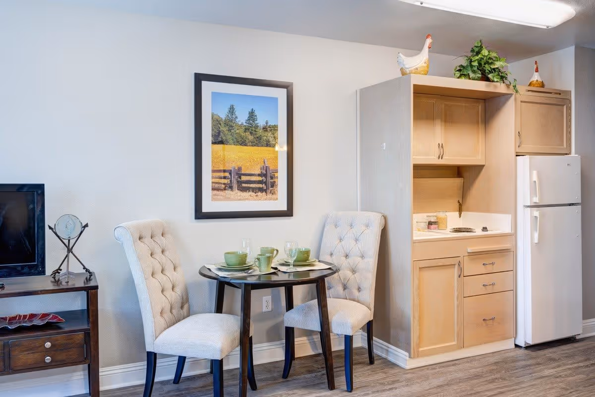 A small dining area with a round dark wooden table set for two with green dishes and glasses. Two beige tufted chairs are positioned at the table. To the right is a compact kitchenette with light wood cabinets, a white refrigerator, and a small countertop with a stove. On top of the cabinets are decorative items including a ceramic chicken and a green plant. On the left side, there is a dark wooden TV stand with a flat-screen TV and a decorative clock. A framed picture of a rural landscape with a wooden fence and trees hangs on the wall above the dining table.