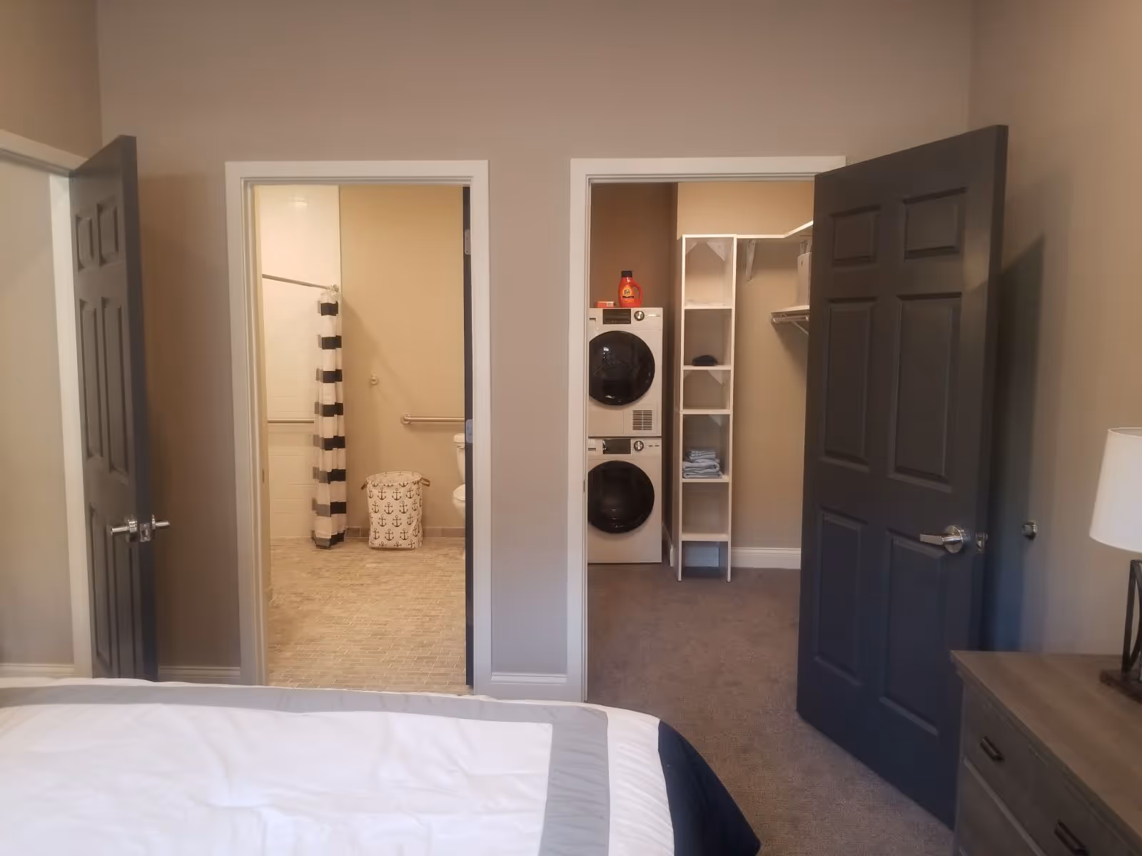 View from a bedroom showing two open doors. The left door leads to a bathroom with a striped shower curtain, grab bars, and a laundry basket with anchor designs. The right door opens to a laundry room with a stacked washer and dryer, shelving with folded towels, and laundry detergent on top. A bedside table with a lamp is partially visible on the right side of the image.