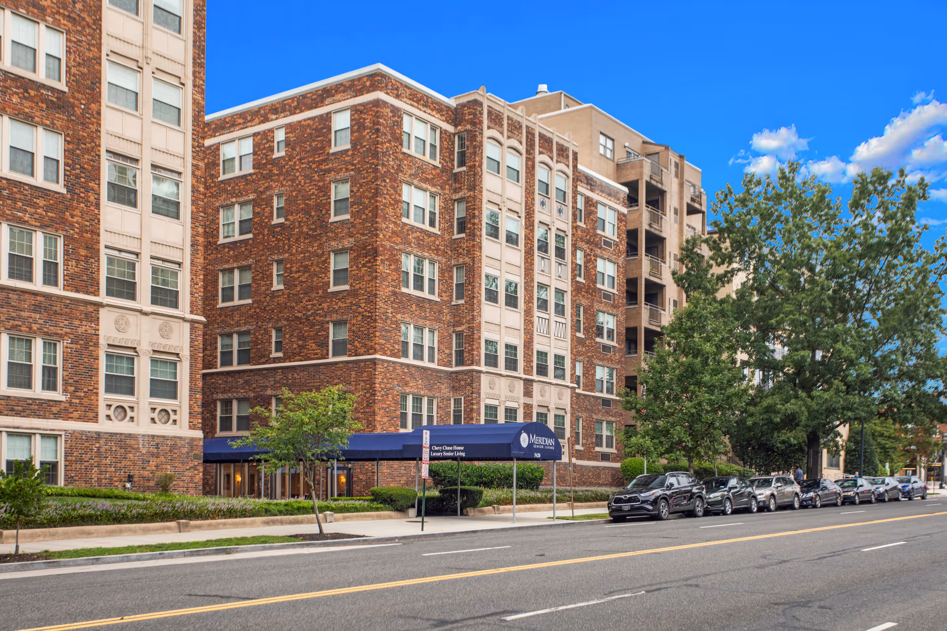 Exterior view of a multi-story brick building with white accents, identified as Chevy Chase House luxury senior living. The building has multiple windows and a blue awning over the entrance. Several cars are parked along the street in front of the building, and there are trees and greenery around the entrance area under a bright blue sky with some clouds.