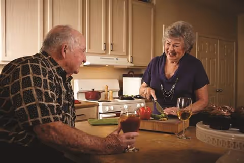An elderly woman is cutting vegetables on a wooden cutting board in a kitchen while an elderly man sits at the counter holding a glass of red wine. The kitchen has light-colored cabinets and a stove in the background. A glass of white wine and a plate of food are also on the counter.