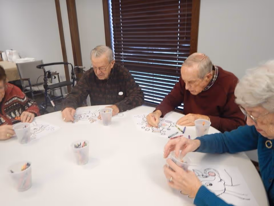Four elderly individuals sitting around a round table engaged in coloring activities with crayons and coloring sheets in a well-lit room with window blinds in the background.