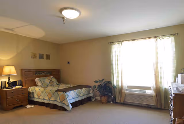 A bedroom in a senior living facility with a single bed covered in a patterned quilt, a wooden nightstand with a lamp and phone, a large window with curtains, an air conditioning unit below the window, a potted plant, and a wooden dresser.