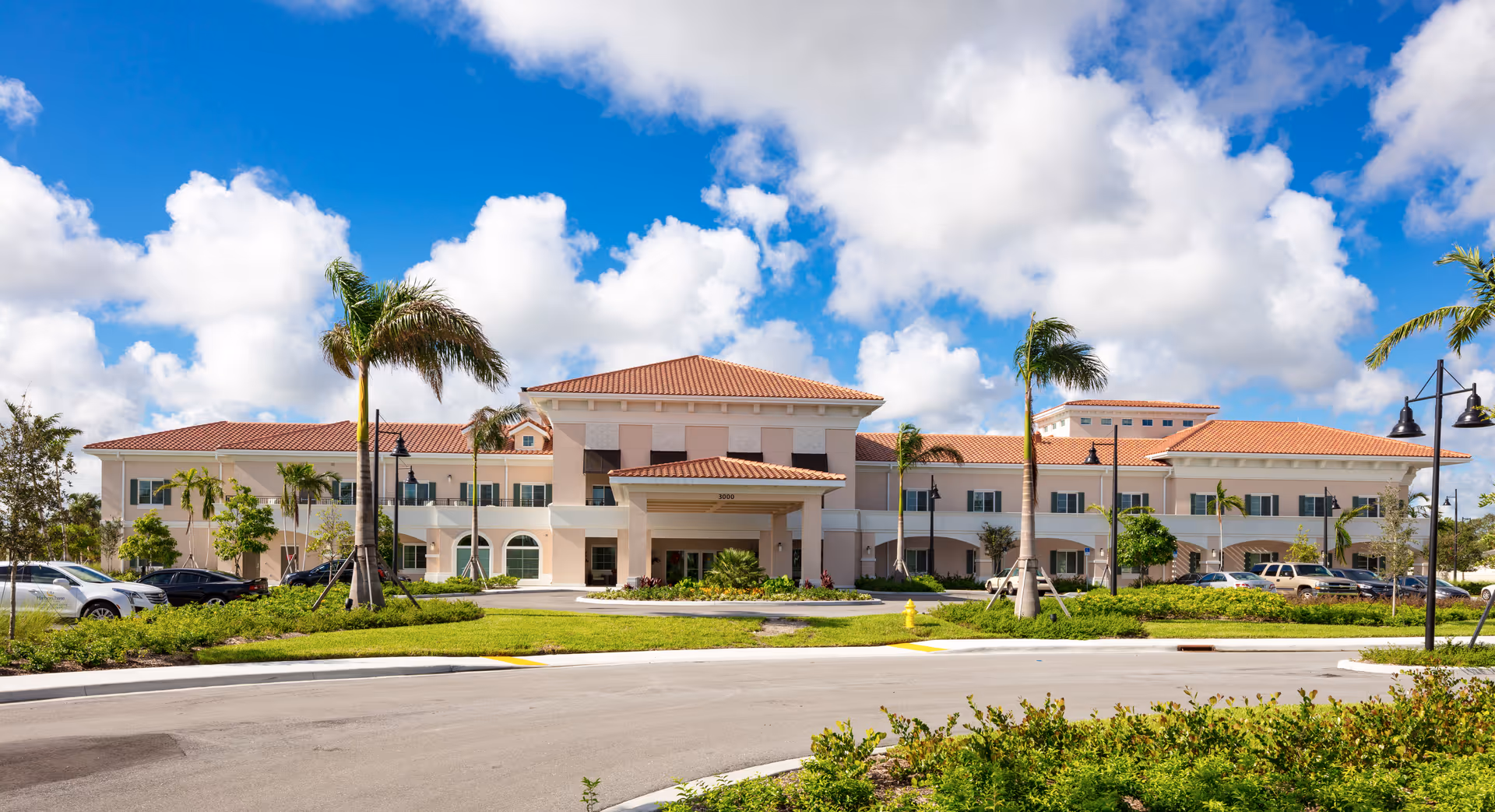 Front exterior view of a large, two-story senior living facility building with a red-tiled roof, palm trees, and a circular driveway under a partly cloudy blue sky.