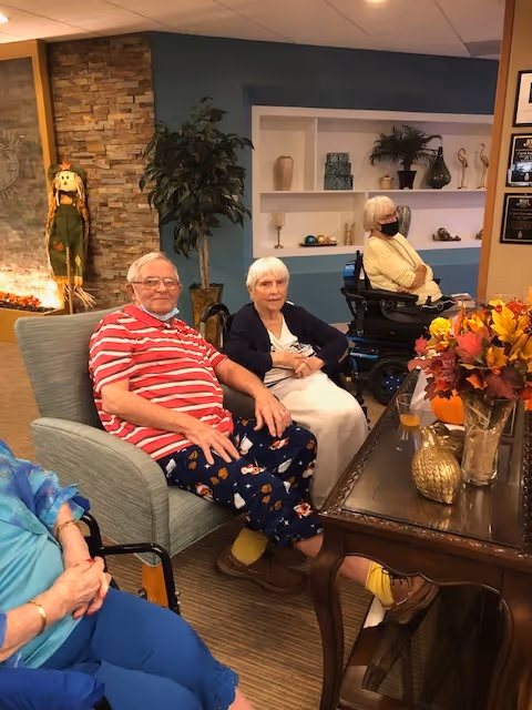A group of elderly people sitting in a cozy living room area with a stone fireplace, decorative shelves, and a table with a fall-themed floral arrangement. One man in a red and white striped shirt and patterned pants is seated in a chair, next to a woman in a dark cardigan and white skirt. Another woman in a wheelchair wearing a yellow top and face mask is in the background.