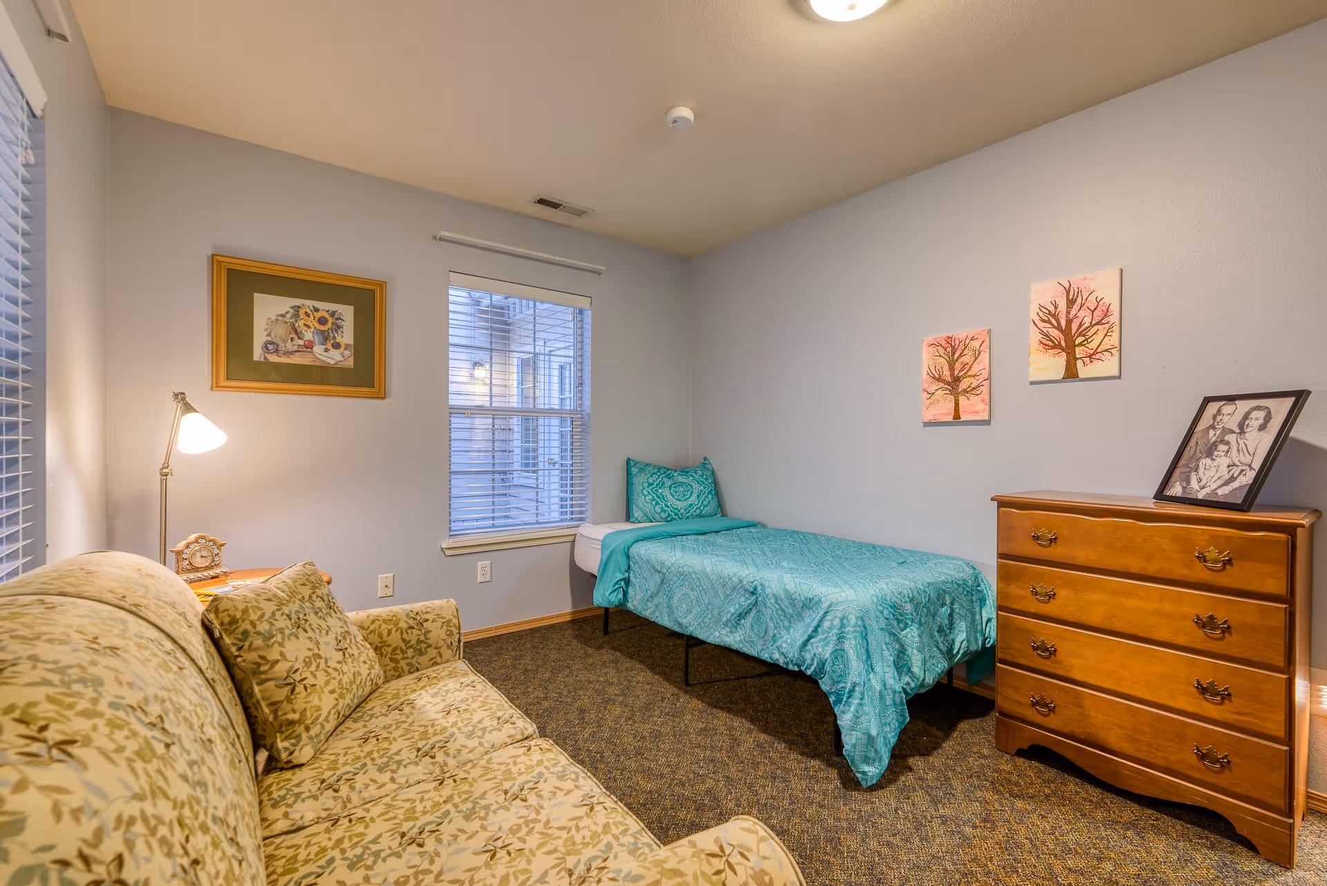 A cozy bedroom with a single bed covered in a teal bedspread, a wooden dresser with a framed black and white photo on top, a floral patterned couch with matching pillows, a floor lamp, and framed artwork on the walls. There is a window with blinds letting in natural light.