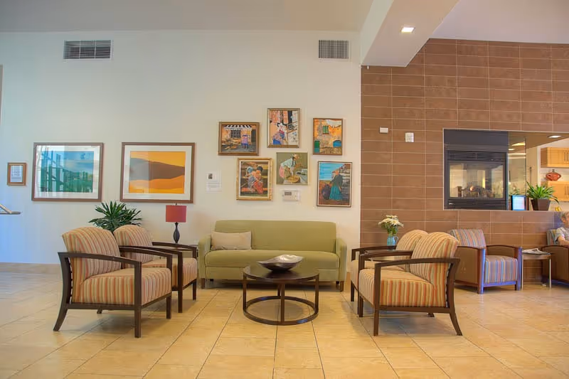 A cozy senior living facility common area with a green sofa, four striped armchairs arranged around a circular coffee table, and a modern fireplace built into a brown tiled wall. The walls are decorated with framed artwork, and there are plants and a small table lamp adding to the warm ambiance.