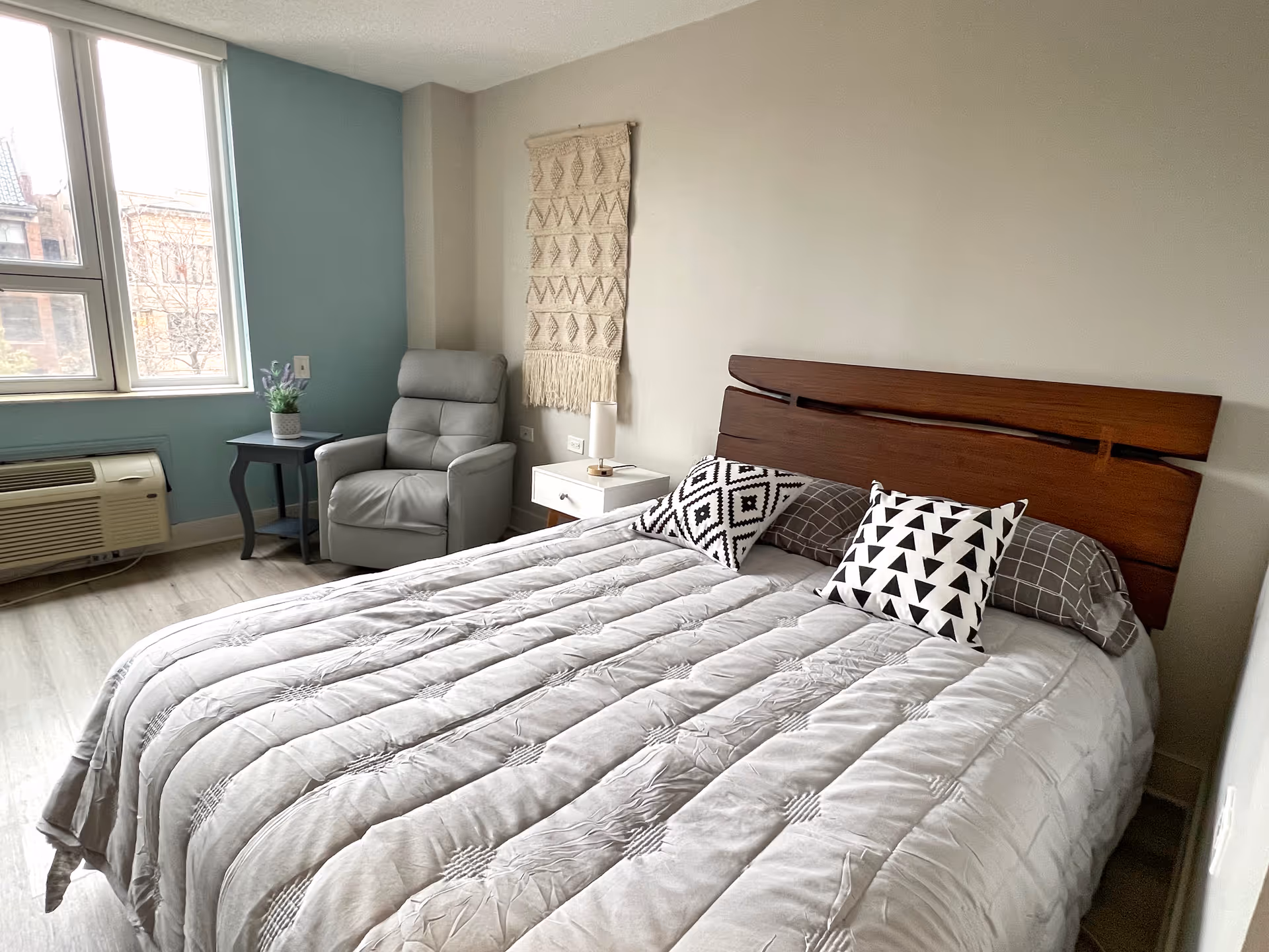 A neatly made bed with a wooden headboard and three decorative pillows in a senior living community bedroom. Next to the bed is a white nightstand with a small lamp. A gray armchair and a small black side table with a potted plant are positioned near a window with a view of buildings outside. The walls are painted in light colors, with one accent wall in teal and a woven wall hanging above the nightstand.