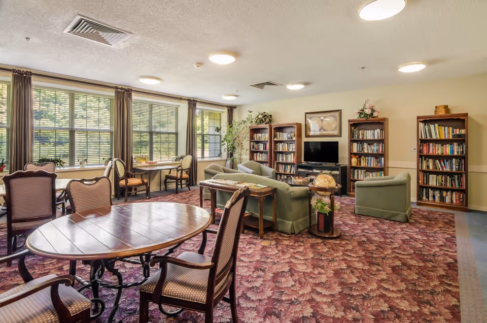 Bright common room in a senior living facility featuring round dining tables, upholstered armchairs, sofas, bookshelves, and a TV.