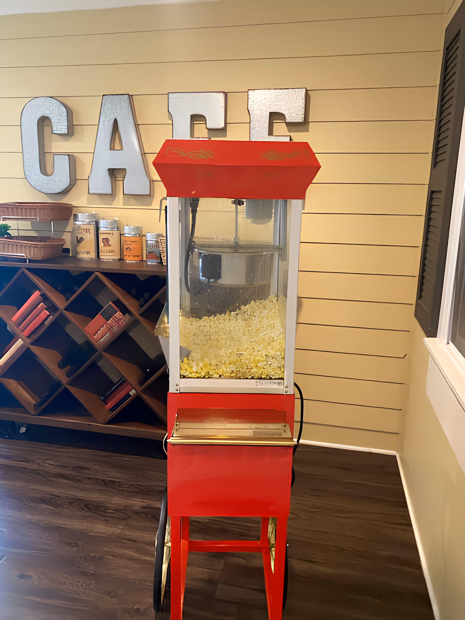 A red vintage-style popcorn machine filled with popcorn is placed on a wooden floor in front of a beige shiplap wall. Behind the machine, there is a wooden wine rack with bottles and containers on top. Large metallic letters spelling 'CAFE' are mounted on the wall above the wine rack.