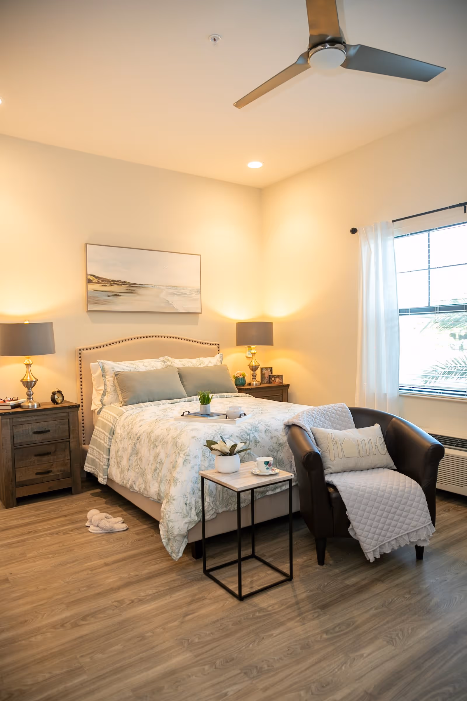 A cozy bedroom with a neatly made bed featuring floral bedding and multiple pillows. On either side of the bed are wooden nightstands with lamps and decorative items. A small side table with a plant and a cup is placed next to a dark leather armchair draped with a light quilt and a pillow. The room has wood flooring, a ceiling fan, and a window with white curtains allowing natural light in.