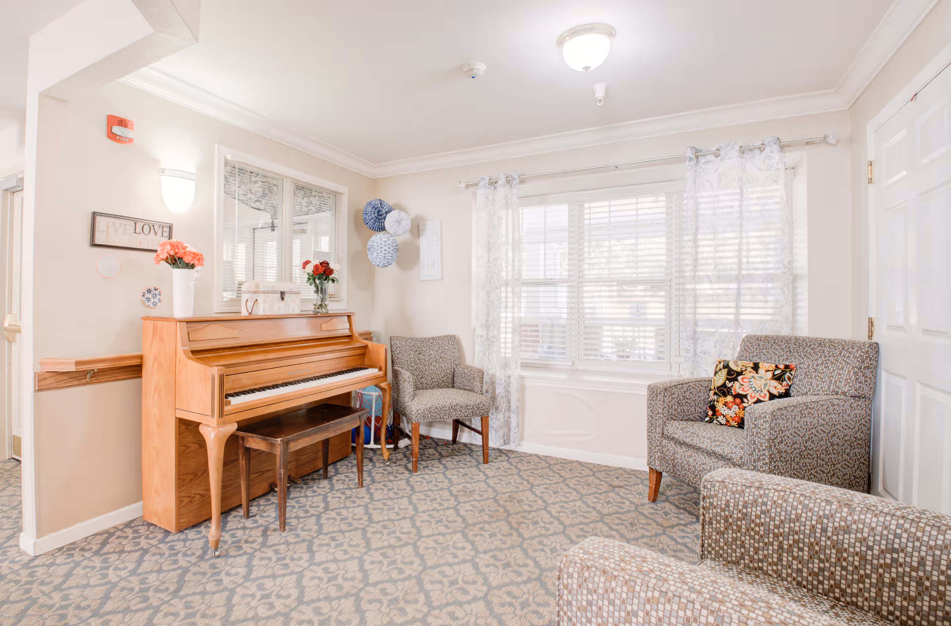 A cozy sitting area in a memory care facility featuring a wooden upright piano with a bench, two patterned armchairs, and a window with sheer curtains letting in natural light. The walls are decorated with a 'Live Love' sign, paper lanterns, and a small framed picture. The carpet has a floral pattern and the room is softly lit with wall and ceiling lights.