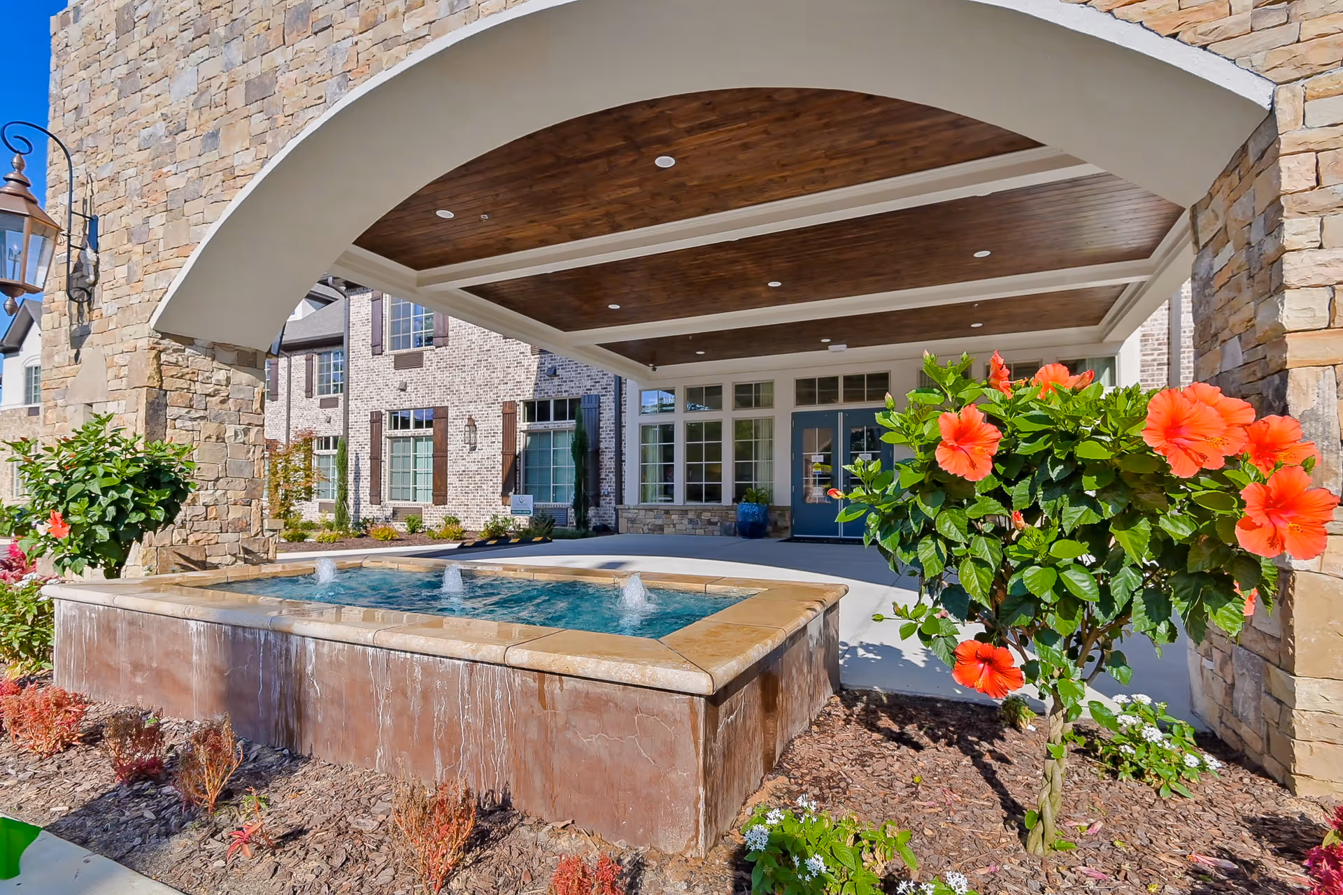 Entrance area of Orchard at Brookhaven featuring a covered stone archway with a wooden ceiling, a rectangular water fountain with three small water jets, and vibrant orange hibiscus flowers in the foreground. The building exterior is made of light-colored brick with large windows and blue double doors.