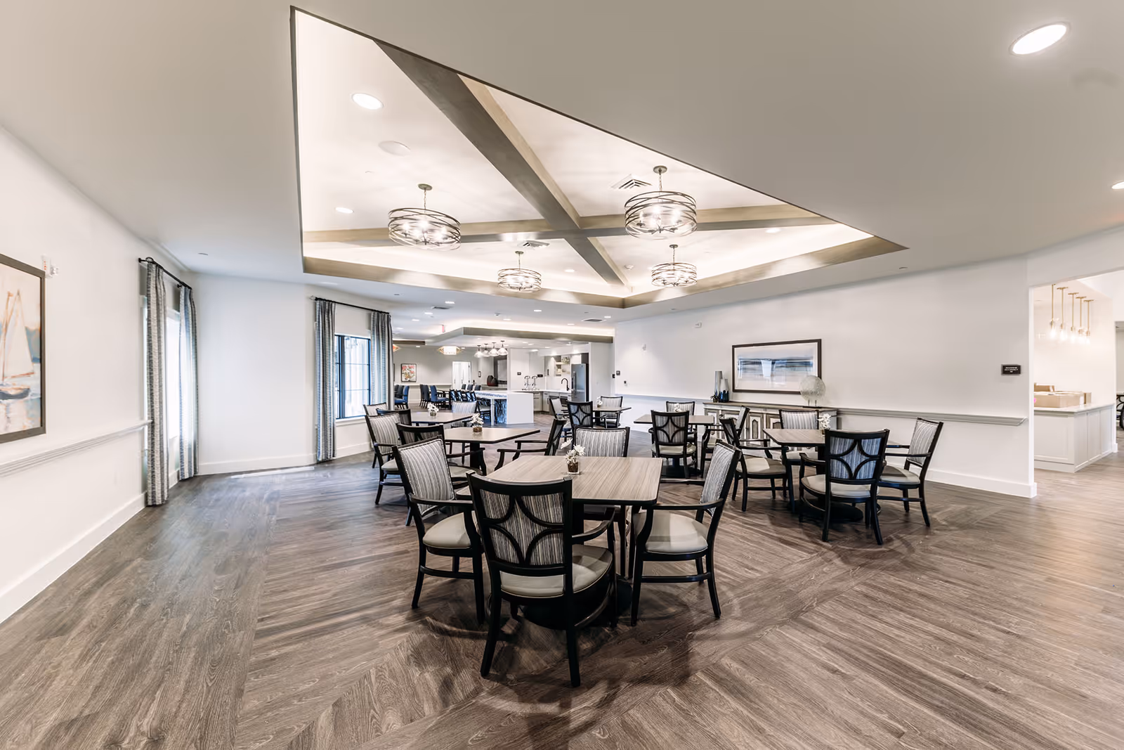 Bright, spacious communal dining area with multiple round tables and chairs, decorative ceiling lights, and a serving area in the background.