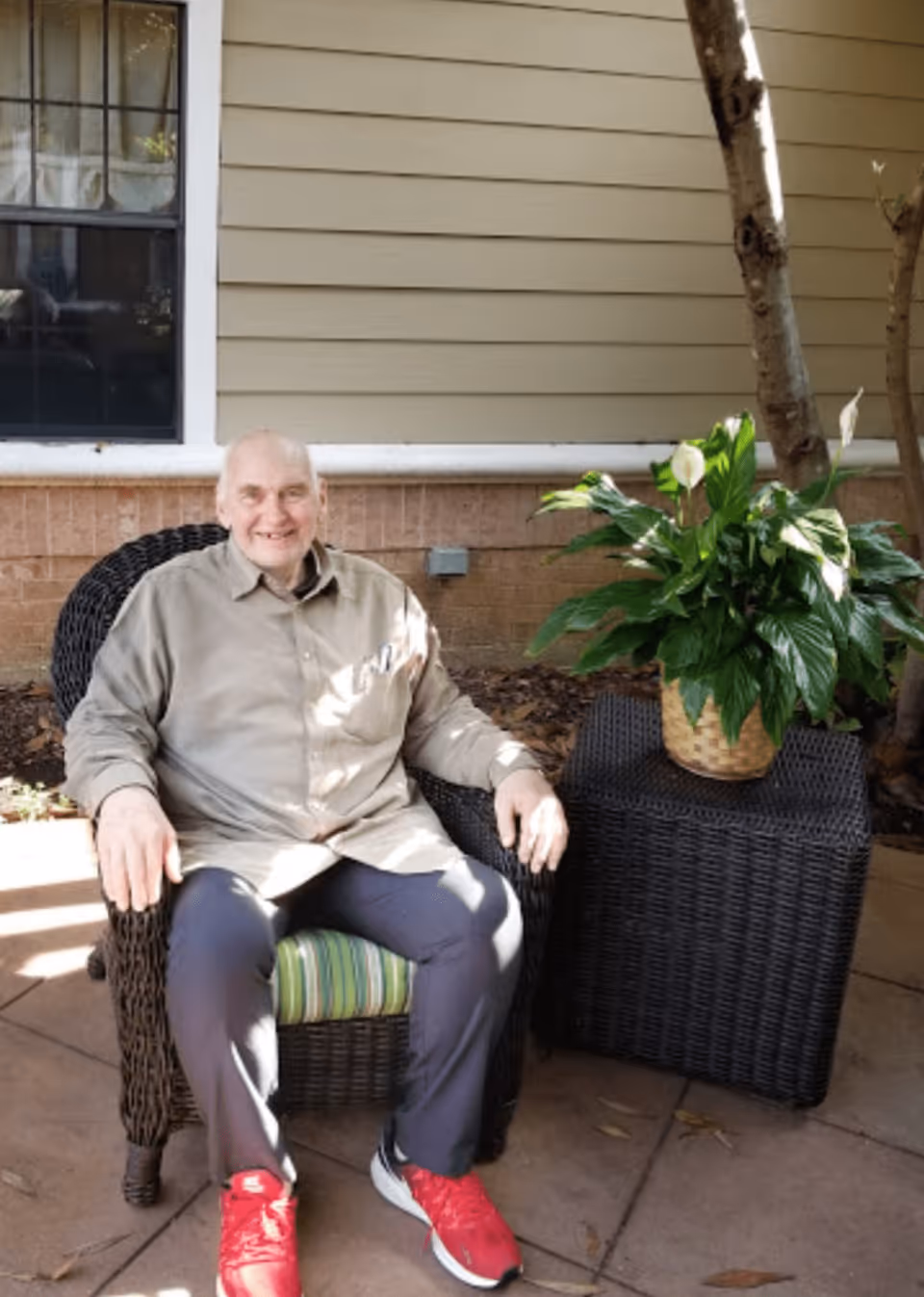 An elderly man with a beige shirt and red sneakers sitting on a wicker chair outside next to a wicker table with a potted plant, in front of a building with beige siding and a window.