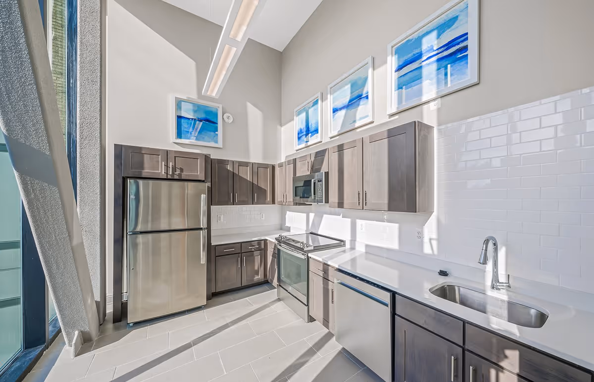Bright modern kitchen with stainless steel refrigerator, stove, microwave, dishwasher, and sink. The kitchen features dark wood cabinets, white countertops, white subway tile backsplash, and three blue abstract paintings on the wall above the cabinets. Large windows allow natural light to fill the space.