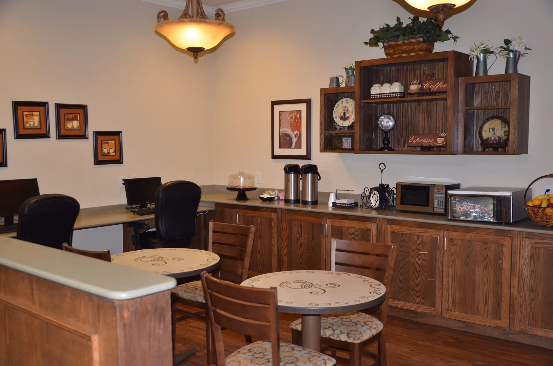 A cozy common area with two round tables and wooden chairs with patterned cushions. There are wooden cabinets along the wall with a microwave, a toaster oven, coffee dispensers, and decorative items including a clock and framed coffee-themed pictures. Two computer workstations with black chairs are visible in the corner. The room has warm lighting from a ceiling fixture.
