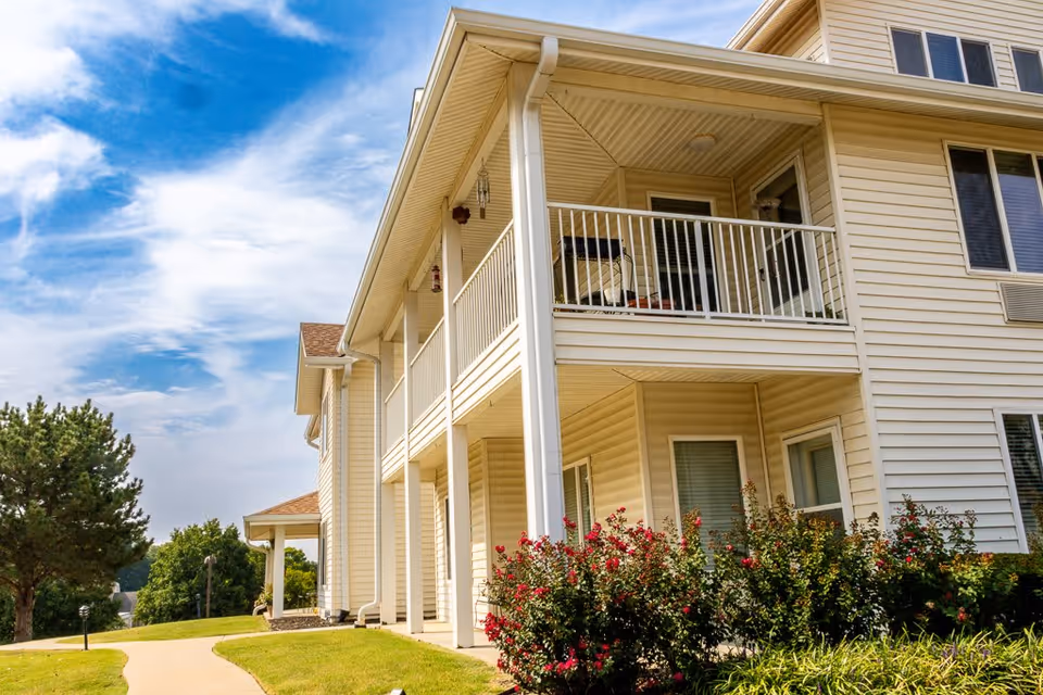 Exterior view of a two-story beige building with a balcony on the upper floor and a covered porch area below. There are bushes with red flowers in front of the building and a paved walkway leading alongside the building. The sky is partly cloudy with blue patches.