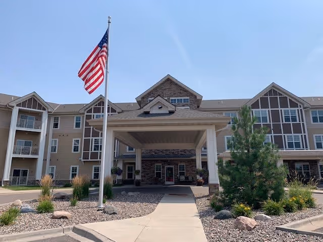 Front exterior view of a three-story senior living facility named Gable Pines with a covered entrance, an American flag on a flagpole, and landscaped areas with rocks and plants.