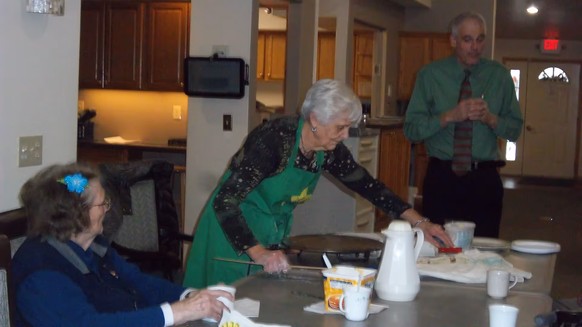 An elderly woman in a green apron prepares food at a table while another seated woman watches and a man stands in the background in a communal dining area.
