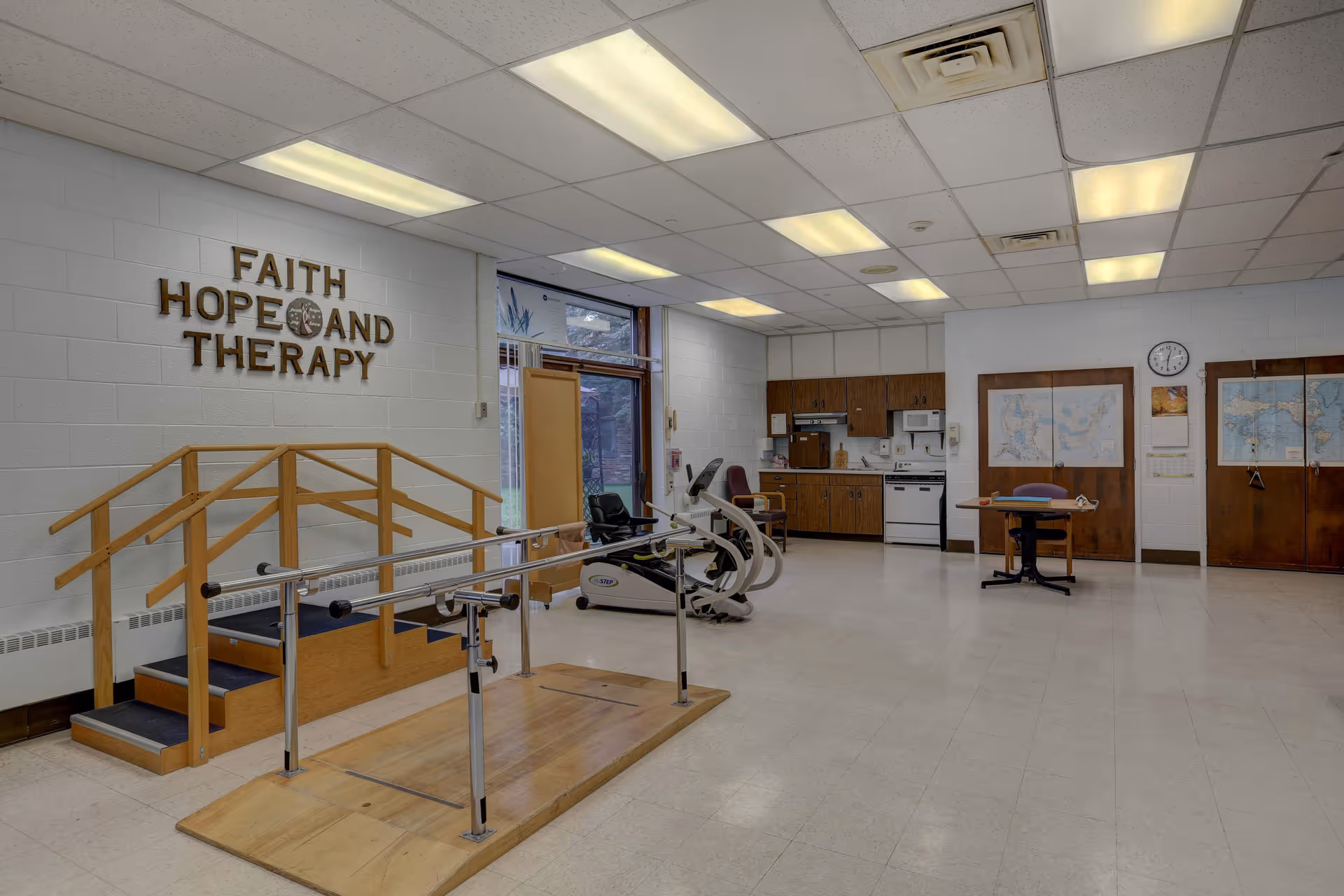 A therapy room with exercise equipment including a stationary bike and parallel bars for walking practice. There is a small wooden staircase with handrails. The walls are white with a sign that reads 'FAITH HOPE AND THERAPY'. The room has a kitchenette area with cabinets, a stove, and a microwave. There is a table with a chair and maps on the wall, along with a clock showing the time.