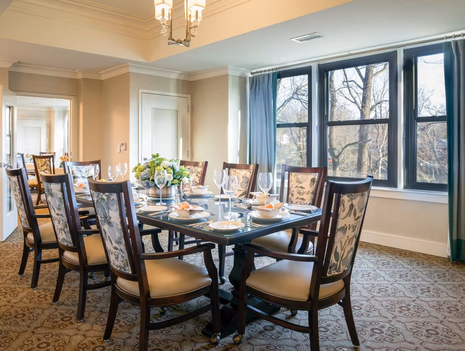 A well-lit dining room with a large rectangular glass-top table set for eight people. The table is adorned with white plates, bowls, napkins, wine glasses, and a centerpiece of green and white flowers. The chairs have wooden frames with floral-patterned upholstery on the backrests and beige cushions. Large windows with blue curtains allow natural light to fill the room, and a chandelier hangs from the ceiling.