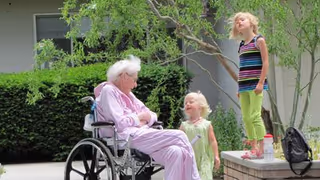 An elderly woman in a wheelchair wearing a pink outfit is outdoors, smiling and interacting with two young girls. One girl is standing on a low brick wall wearing a striped tank top and green pants, while the other girl stands beside the wheelchair wearing a light green dress. There is greenery and a building in the background.