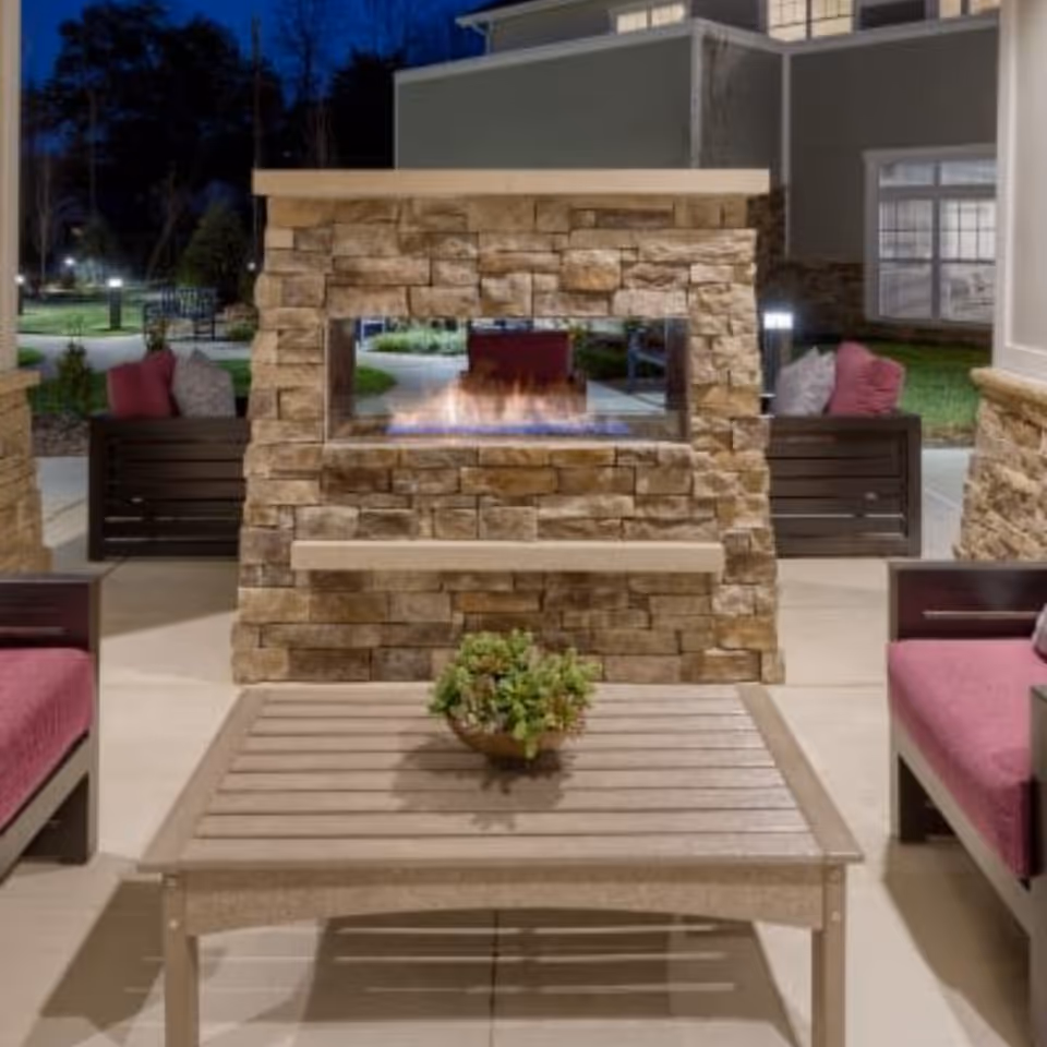 Outdoor seating area at night with a stone fireplace in the center, surrounded by cushioned chairs with red and gray pillows, and a wooden coffee table with a small plant on it.