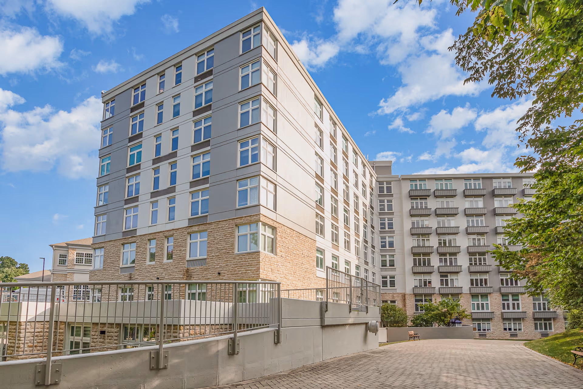 Exterior view of a multi-story senior living facility building with numerous windows and balconies, a paved walkway, and trees on the right side under a partly cloudy blue sky.