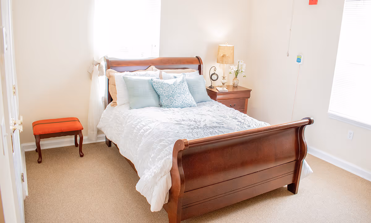 A bright bedroom with a wooden sleigh bed made with white bedding and multiple pillows in shades of blue and beige. There is a wooden nightstand with a lamp, clock, and a small vase with flowers. A small red cushioned bench is placed near the door, and the room has beige carpet and white walls with two windows letting in natural light.