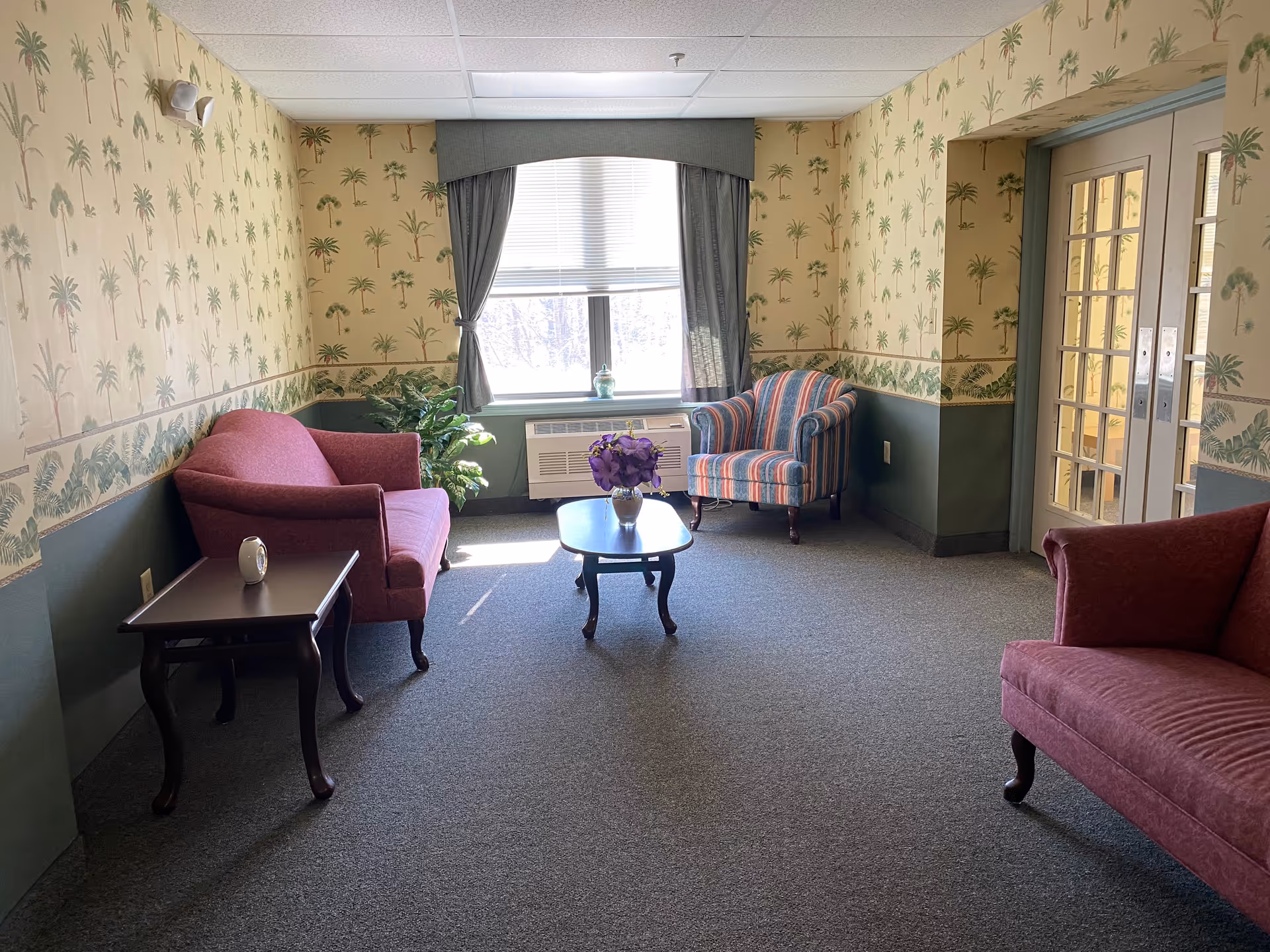 A cozy sitting area in a senior living facility with two red upholstered loveseats, a striped armchair, a small wooden coffee table with a vase of purple flowers, and a side table with a small clock. The walls have palm tree patterned wallpaper with a green lower border, and a window with gray curtains lets in natural light.