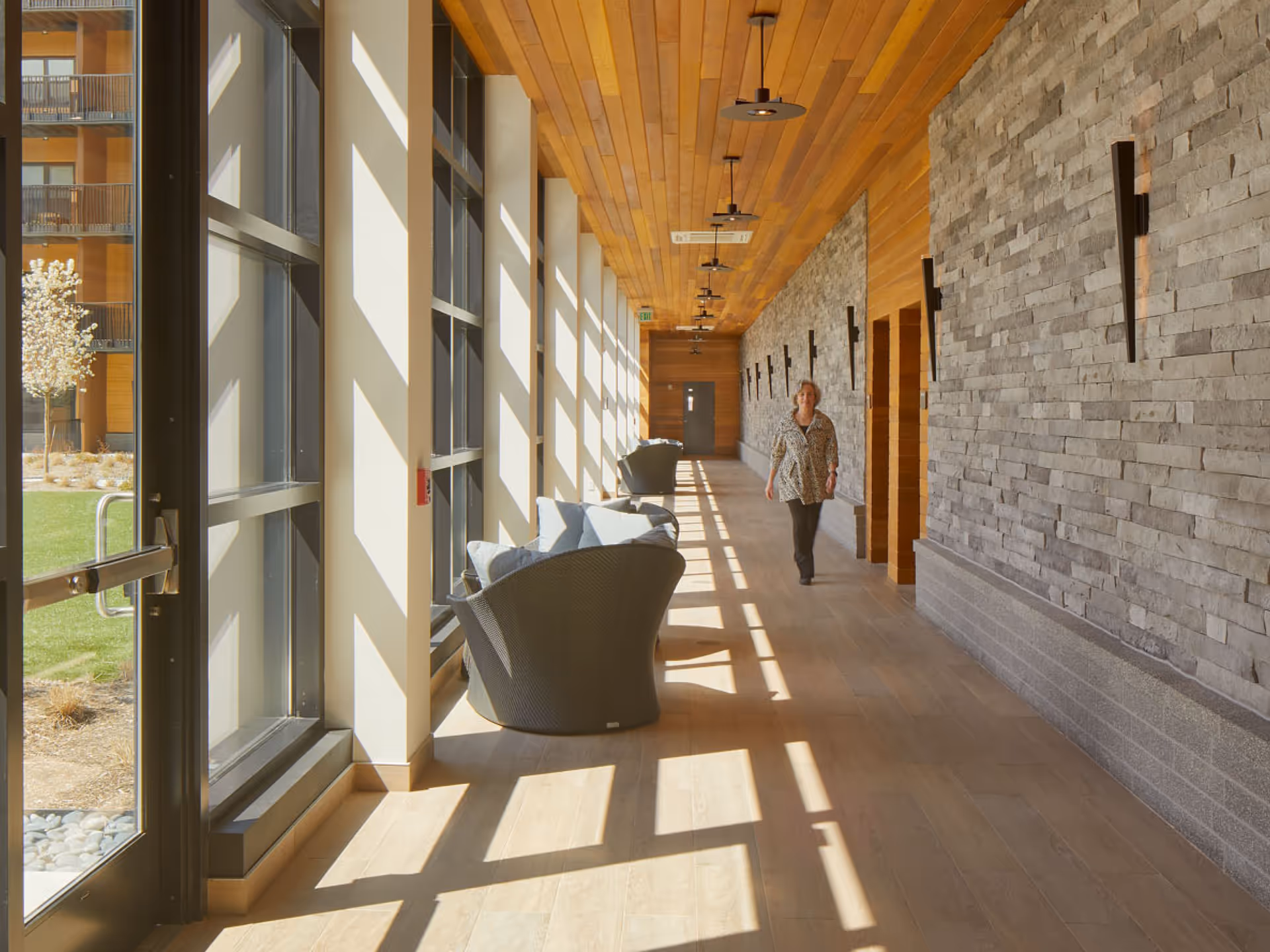 A sunlit corridor with large windows on the left side casting shadows on the wooden floor. There are comfortable wicker chairs with cushions along the window side. The right wall is made of textured stone with modern wall sconces. A woman is walking down the corridor towards the camera. Outside the windows, a green lawn and a tree are visible.