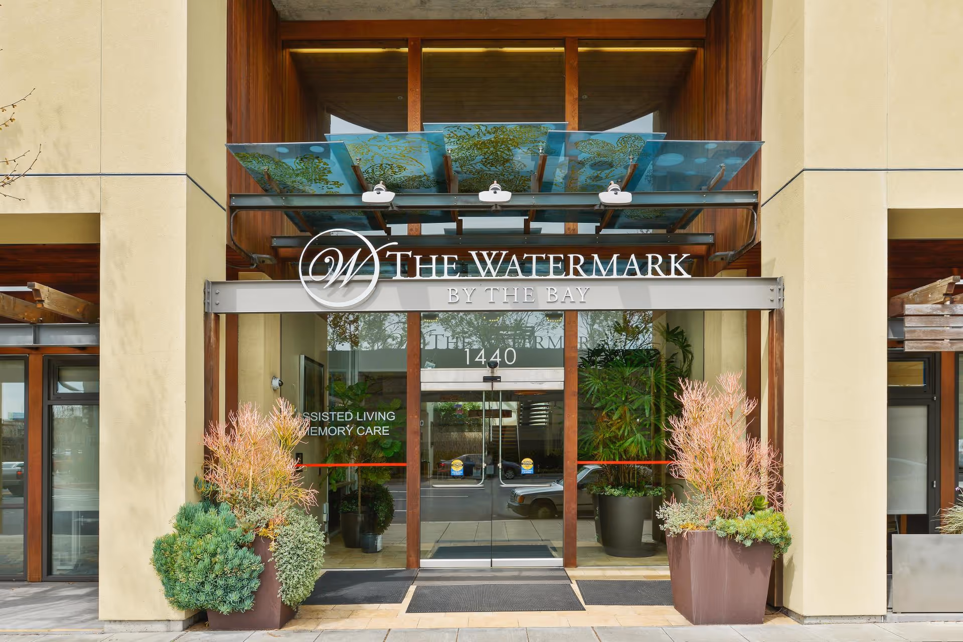 Entrance to The Watermark by the Bay senior living facility with glass doors, potted plants on either side, and a decorative glass canopy above the door.