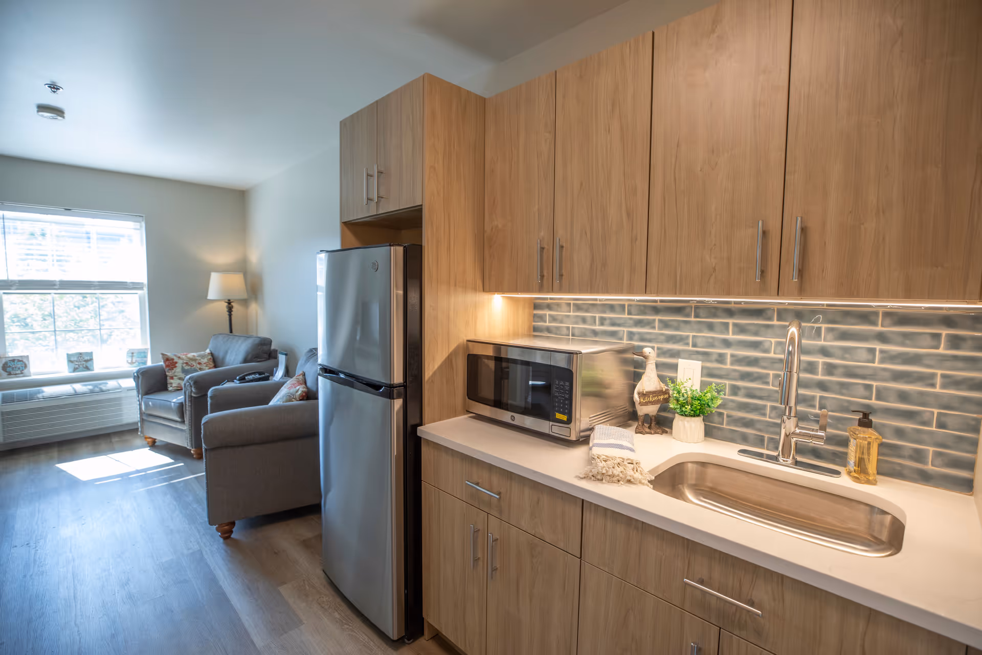 Compact open-plan kitchen and sitting area with wood cabinets, stainless refrigerator, microwave, sink, and two gray armchairs by a window.