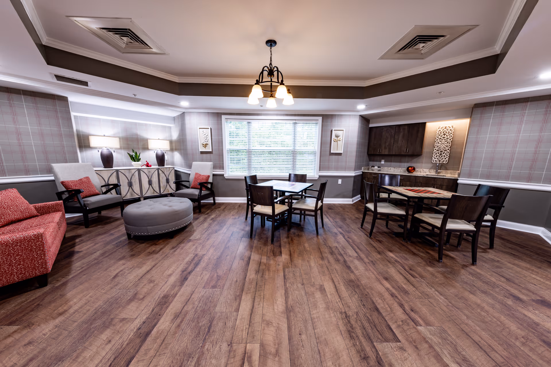Bright communal dining and sitting room with wood flooring, armchairs, a round ottoman, and several dining tables.