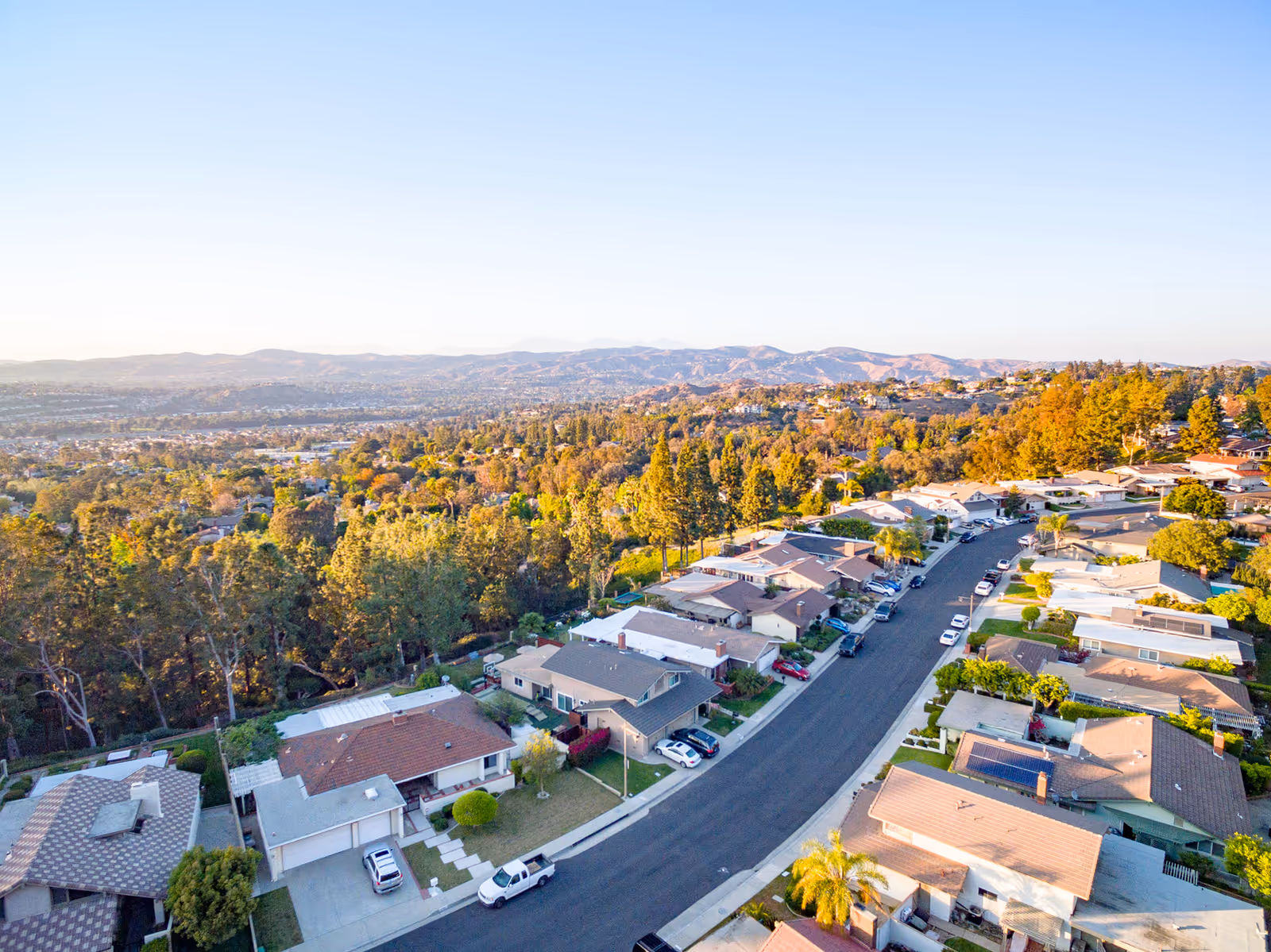 Aerial view of a suburban neighborhood with single-family homes, tree-lined streets, and a backdrop of rolling hills under a clear blue sky.
