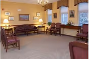 A bright communal sitting room with upholstered chairs and a sofa, lamps, framed artwork, and windows with valances.