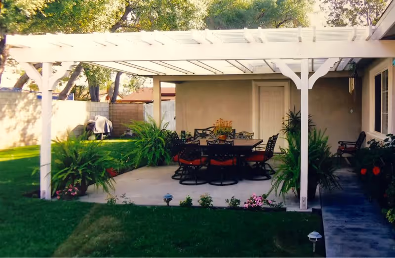 Outdoor patio area with a white pergola covering a dining table surrounded by eight chairs with red cushions. The patio is surrounded by green grass, potted plants, and flowers. There is a beige wall with a door in the background and trees providing shade.